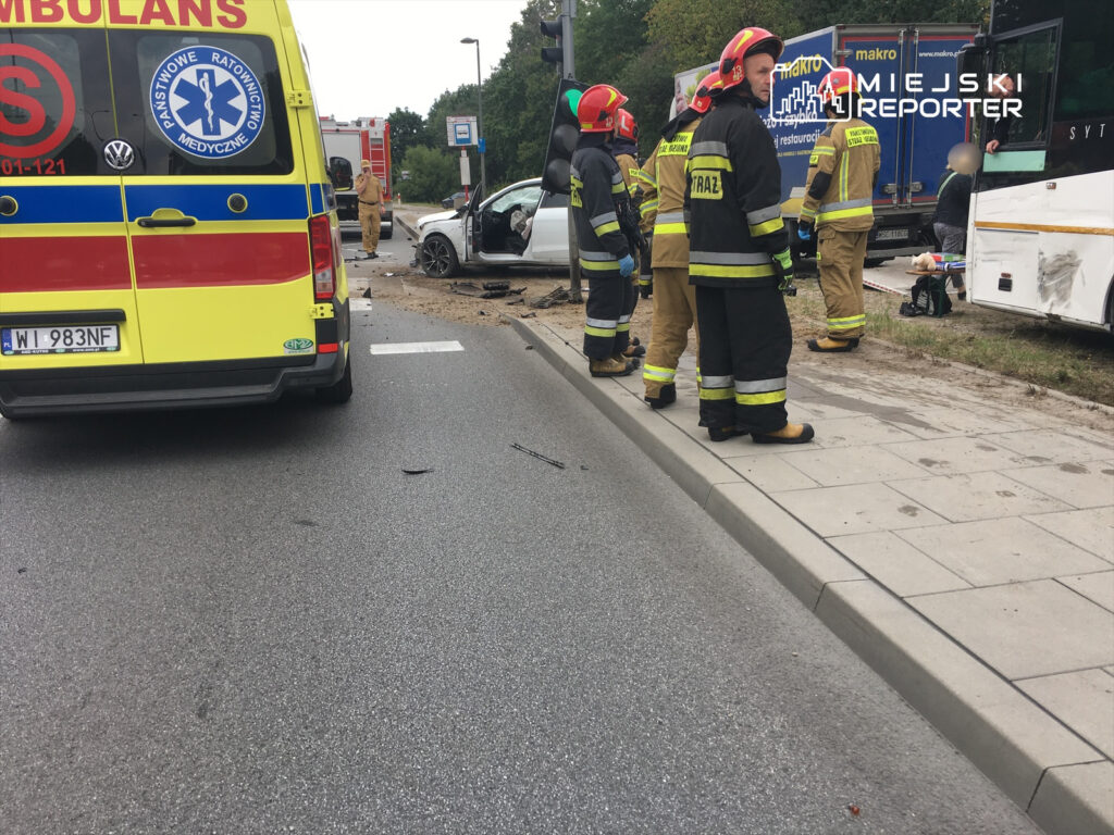 Grupa strażaków w mundurach omawia sytuację obok uszkodzonego samochodu osobowego na drodze. Obok stoi ambulans.