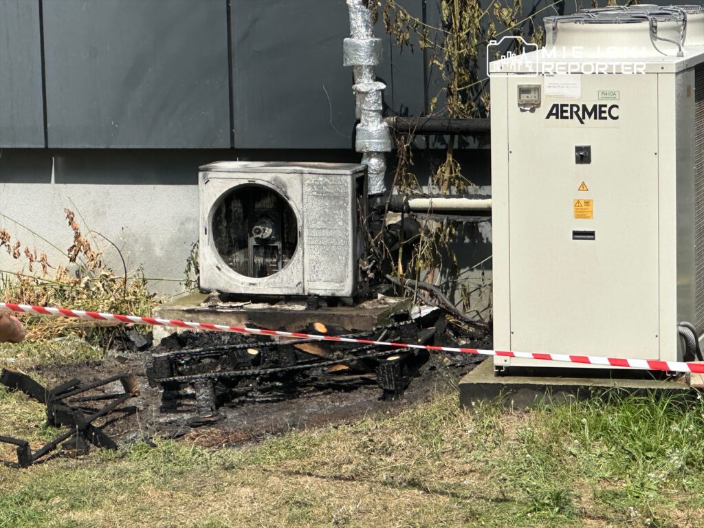 Uszkodzona jednostka klimatyzacyjna obok spalonego podłoża, zabezpieczona taśmą ostrzegawczą.