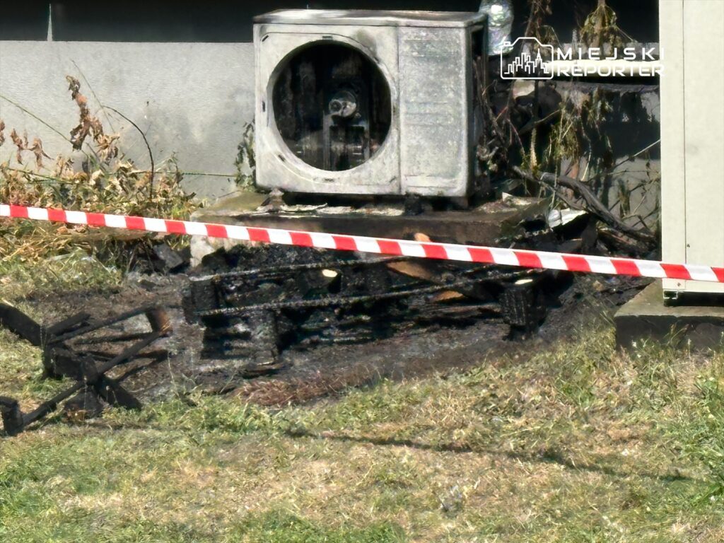 Spalona instalacja klimatyzacyjna otoczona taśmą ostrzegawczą na trawie w pobliżu budynku.