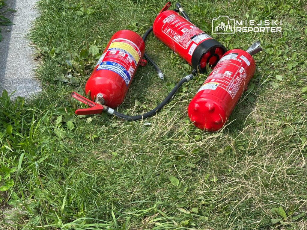 Trzy gaśnice w czerwonym kolorze leżą na trawie obok chodnika.