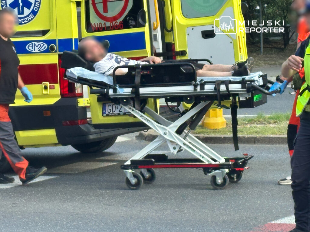 Mężczyzna leżący na noszach, transportowany przez ratowników medycznych obok karetki na ulicy.