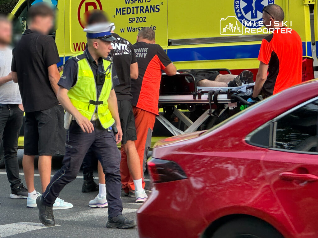 Policjant w kamizelce odblaskowej przeprowadza czynności na miejscu zdarzenia, podczas gdy ratownicy medyczni transportują poszkodowanego na noszach. W tle widoczna karetka pogotowia.