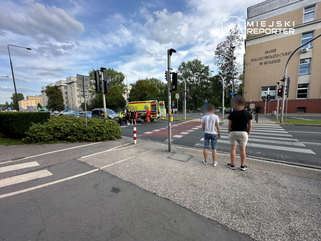 Dwaj mężczyźni stoją na przejściu dla pieszych, obserwując działania służb ratunkowych przy ulicy, obok nowoczesnego budynku.