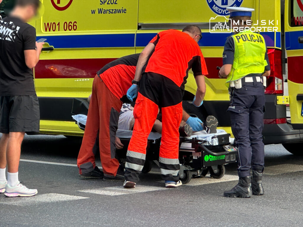 Ratownicy medyczni udzielają pomocy mężczyźnie leżącemu na noszach obok karetki, a policjant nadzoruje sytuację.