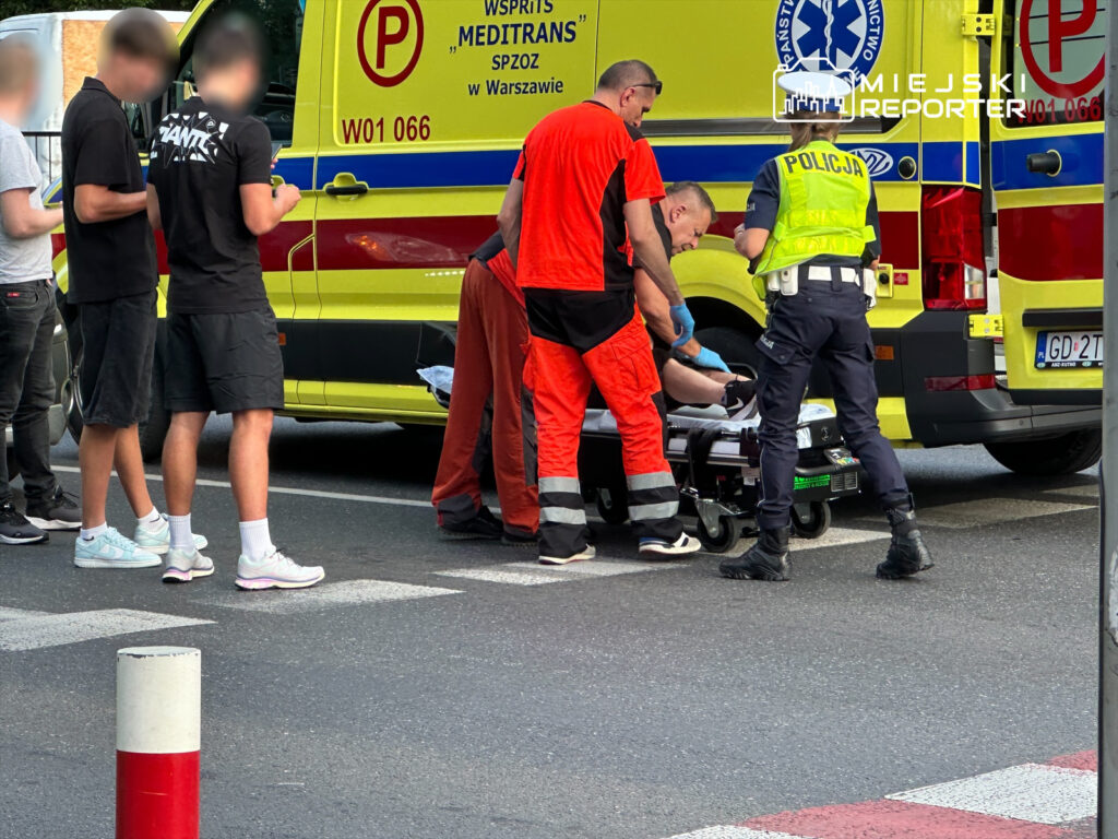 Grupa osób obserwuje ratowników medycznych udzielających pomocy poszkodowanemu przy ambulansie.