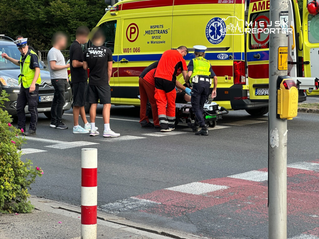 Grupa osób wokół ratowników medycznych udzielających pomocy poszkodowanemu na ulicy, obok karetki i radiowozu.