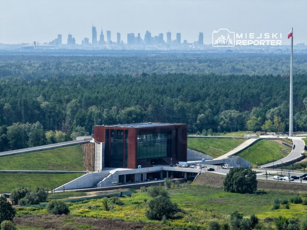 Nowoczesny budynek z przeszkloną fasadą w otoczeniu lasu, w tle panorama miejskiego krajobrazu.