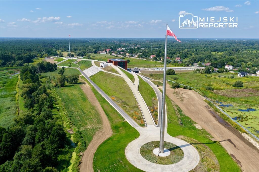 Wysoka flaga powiewająca na maszcie obok nowoczesnego budynku, otoczonego zielonym terenem i ścieżkami.