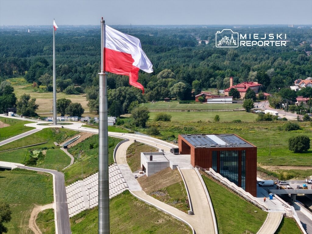 Flaga Polski powiewa na masztach w otoczeniu nowoczesnego budynku z przeszkloną fasadą oraz zielonych terenów.
