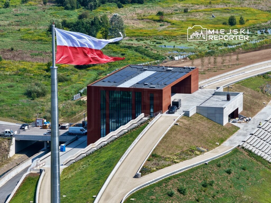 Polska flaga powiewająca na maszcie obok nowoczesnego budynku z przeszkloną fasadą i panelami słonecznymi.