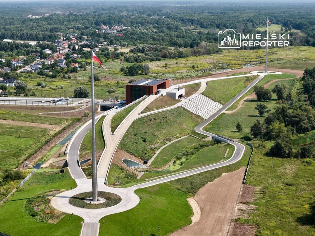 Nowoczesny obiekt z przeszkloną fasadą otoczony zielenią, z flagą powiewającą na maszcie w centralnym punkcie terenu.