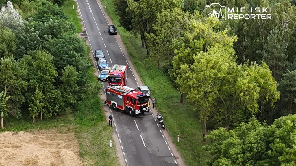 Straż pożarna z wozem bojowym zatrzymała się na drodze, obok stojące pojazdy i zieleń drzew w tle.