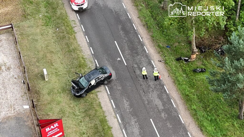 Czarny samochód osobowy z otwartą maską stoi na poboczu drogi, obok dwóch policjantów w kamizelkach odblaskowych.