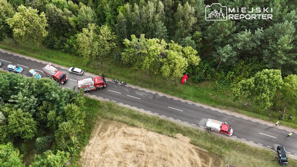 Na drodze, obok lasu, znajdują się dwa wozy strażackie oraz kilka radiowozów policyjnych, a w tle widać zarośla.
