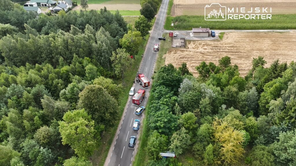 Na drodze w otoczeniu lasu i pól stoją pojazdy straży pożarnej oraz radiowozy, a obok widać zniszczony samochód.
