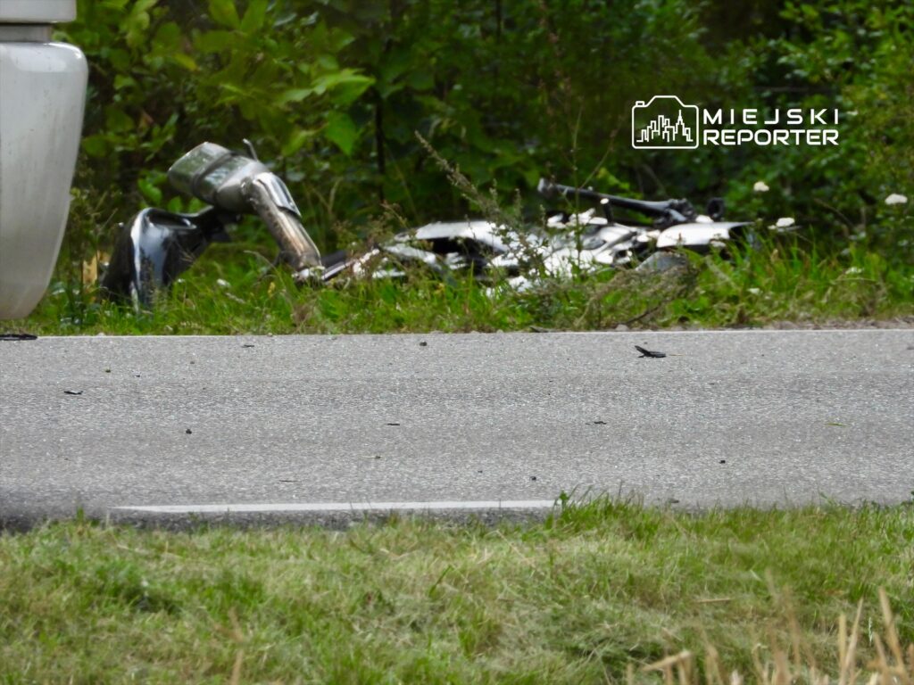 Motocykl leży przewrócony w zaroślach obok drogi, otoczony zielenią i kwiatami.