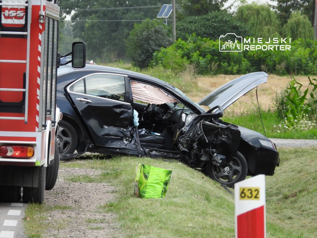 Czarny samochód osobowy z uszkodzoną stroną, zatrzymany na poboczu drogi obok wozu strażackiego.