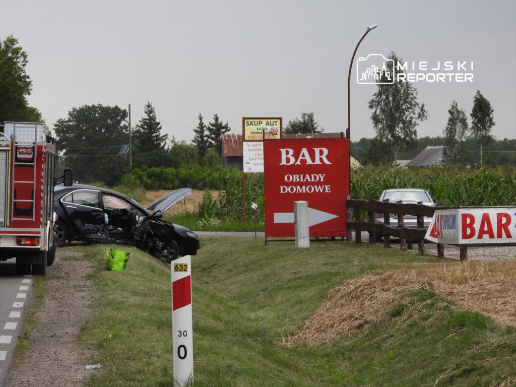Samochód osobowy z uszkodzoną przednią częścią stoi na poboczu drogi, obok wozu strażackiego. W tle widoczny bar z napisem "OBIADY DOMOWE".
