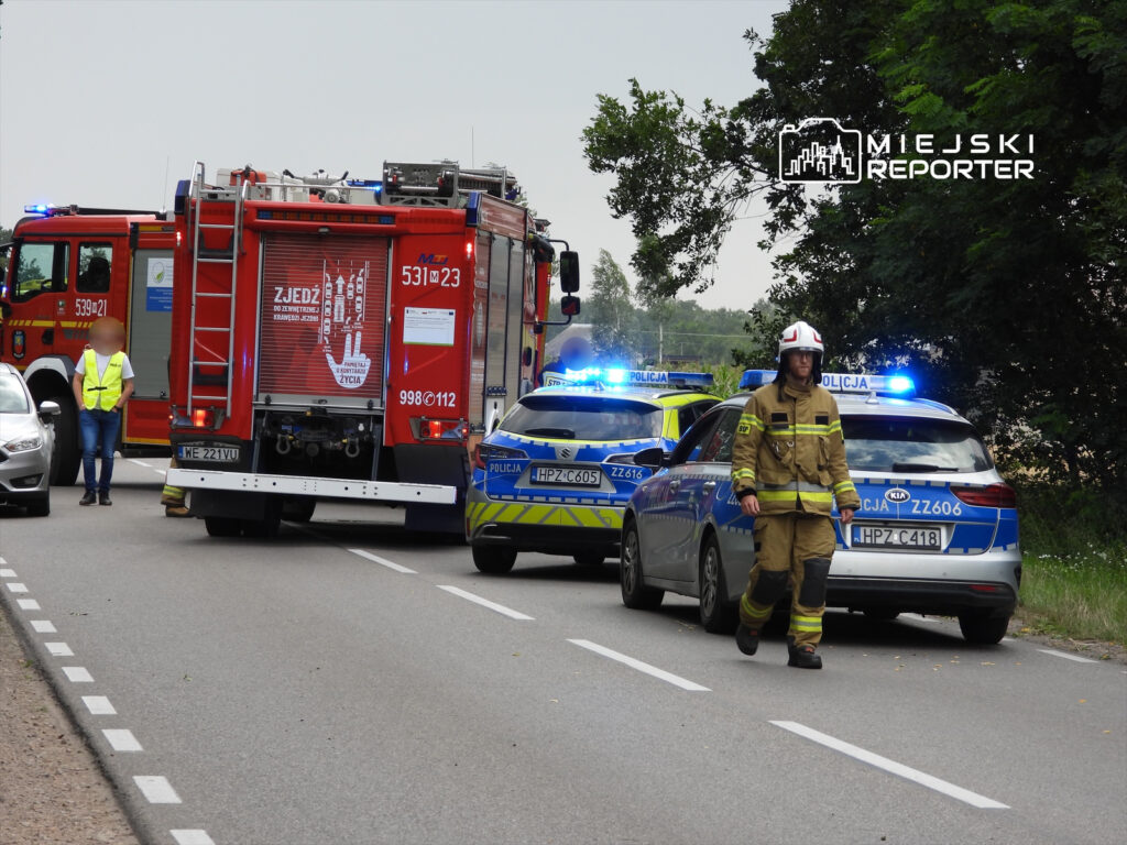Strażak w mundurze kieruje ruchem obok wozu strażackiego i radiowozów policyjnych na drodze.