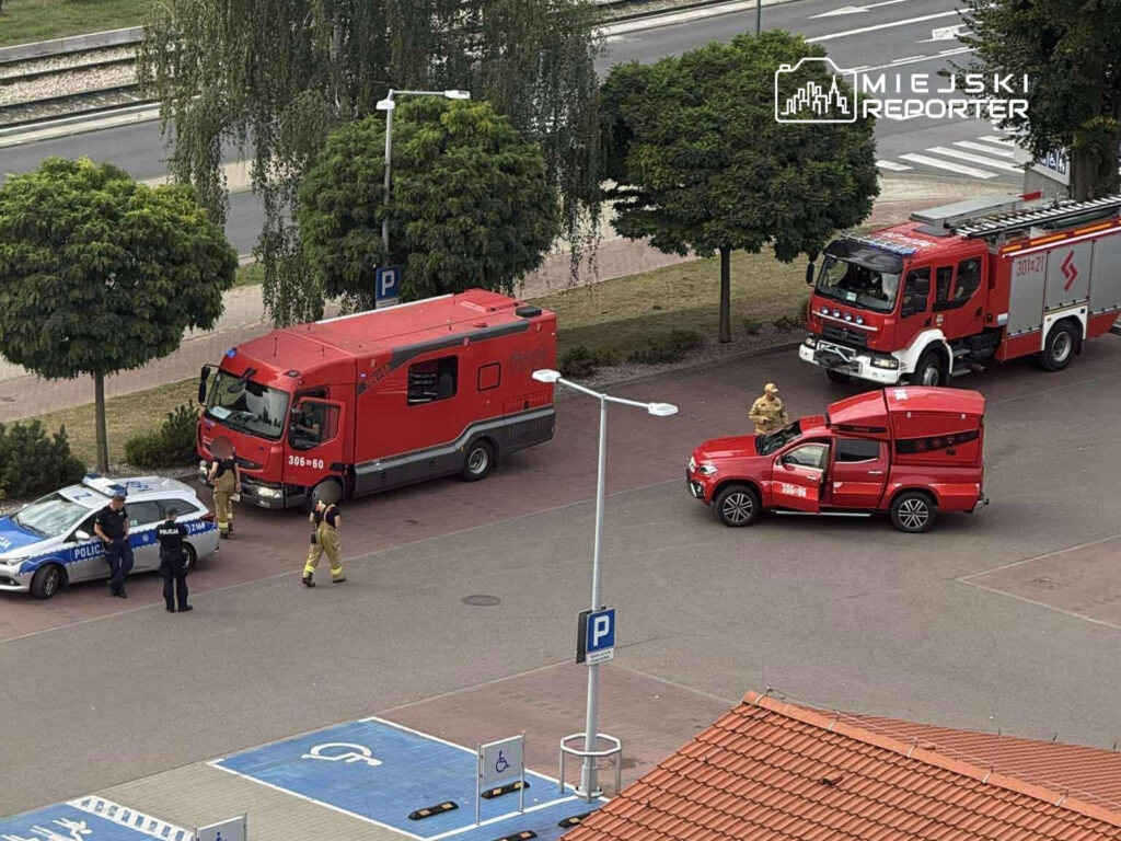 Grupa strażaków oraz policjantów prowadzi działania interwencyjne obok zaparkowanych pojazdów ratunkowych.