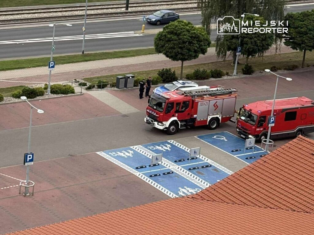 Dwa pojazdy straży pożarnej zaparkowane na parkingu, obok grupy osób rozmawiających w pobliżu.