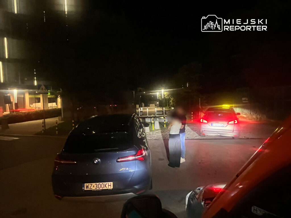 Kobieta i mężczyzna stoją na drodze obok zaparkowanych samochodów w nocy, w tle widać oświetlony budynek.
