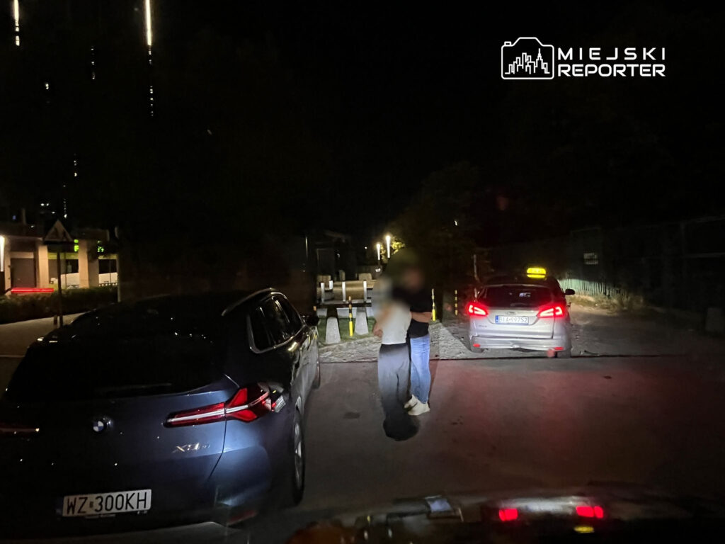 Mężczyzna i kobieta w ciemnym otoczeniu rozmawiają obok dwóch zaparkowanych samochodów.