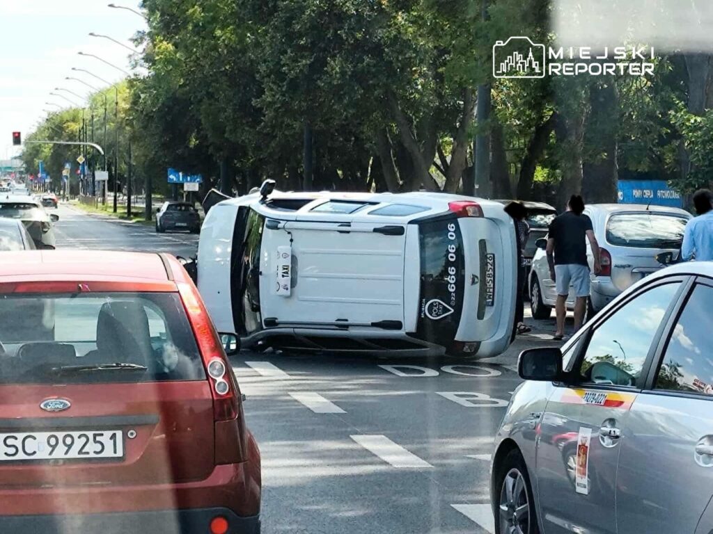Na jezdni leży przewrócony samochód dostawczy, a obok grupka ludzi obserwuje sytuację. W tle widać inne pojazdy i sygnalizację świetlną.