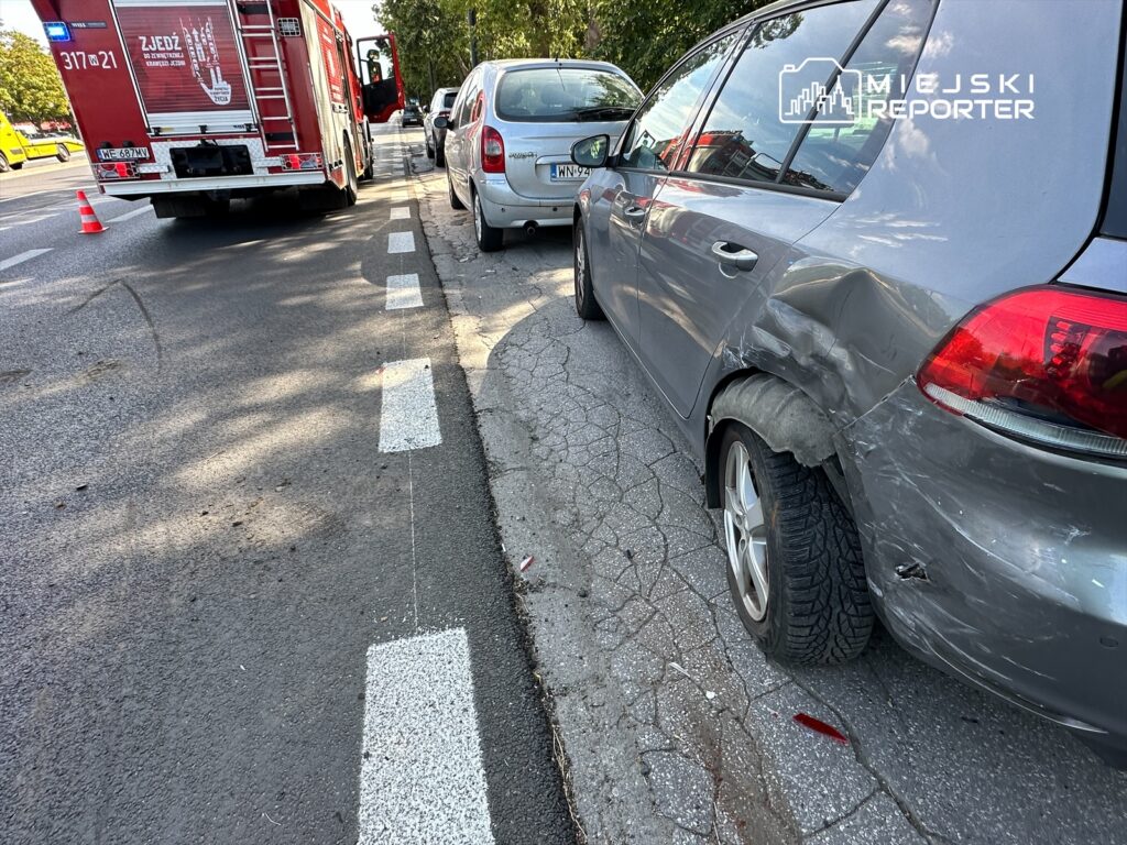 Uszkodzony samochód osobowy zaparkowany na poboczu, obok wozu strażackiego, z widocznymi śladami kolizji.