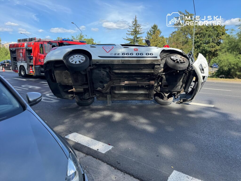 Przewrócone na dach taxi z widocznymi uszkodzeniami, obok straż pożarna oraz inne pojazdy na ruchliwej ulicy.