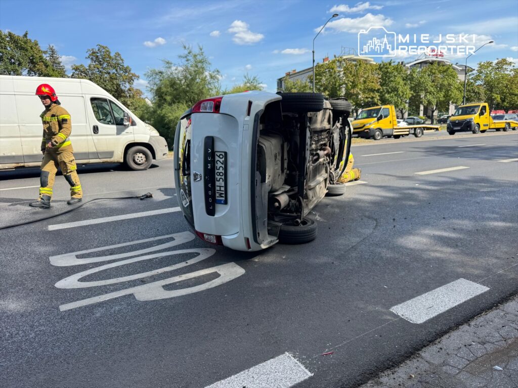 Strażak w hełmie kontroluje przewrócone na bok auto na drodze, obok przejeżdżają inne pojazdy.