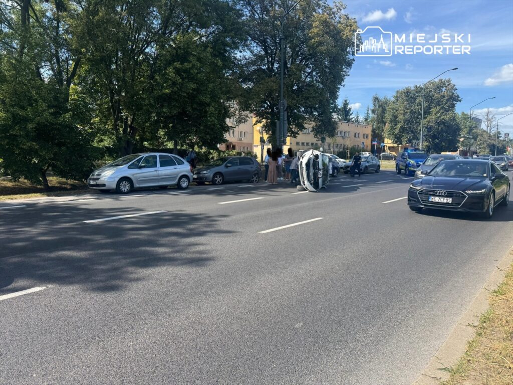 Na jezdni leży przewrócone auto, a obok zgromadził się tłum ludzi. W tle widać inne pojazdy i budynki.