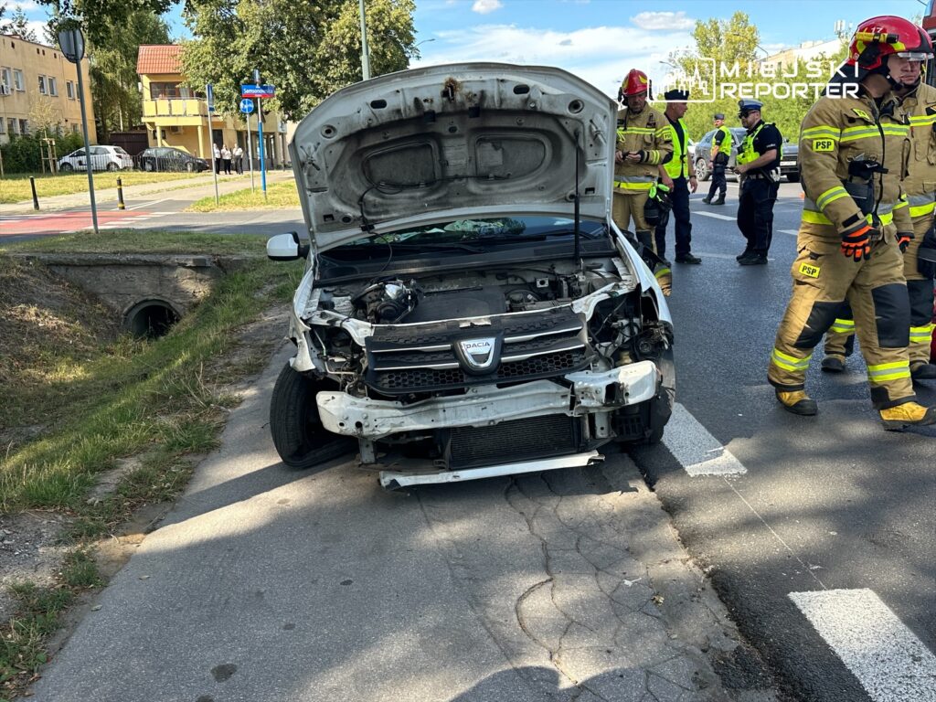 Uszkodzony samochód osobowy na poboczu drogi, obok grupy strażaków i policjantów, w tle budynki mieszkalne.