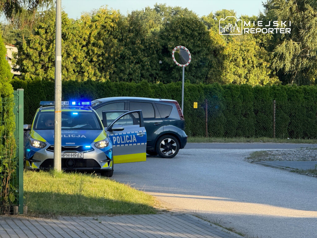 Policyjny radiowóz z włączonymi sygnałami świetlnymi zatrzymany na skrzyżowaniu, obok otwartego samochodu osobowego.