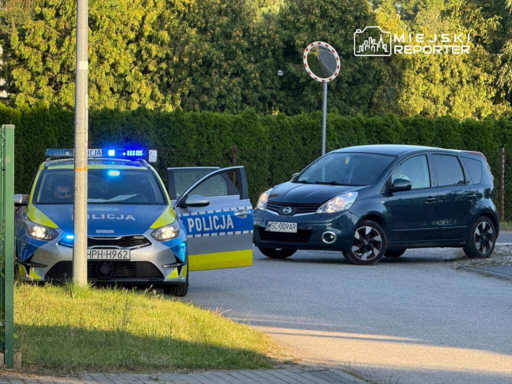 Policyjny radiowóz z włączonymi sygnałami świetlnymi zatrzymany przy drodze obok ciemnoniebieskiego samochodu osobowego.