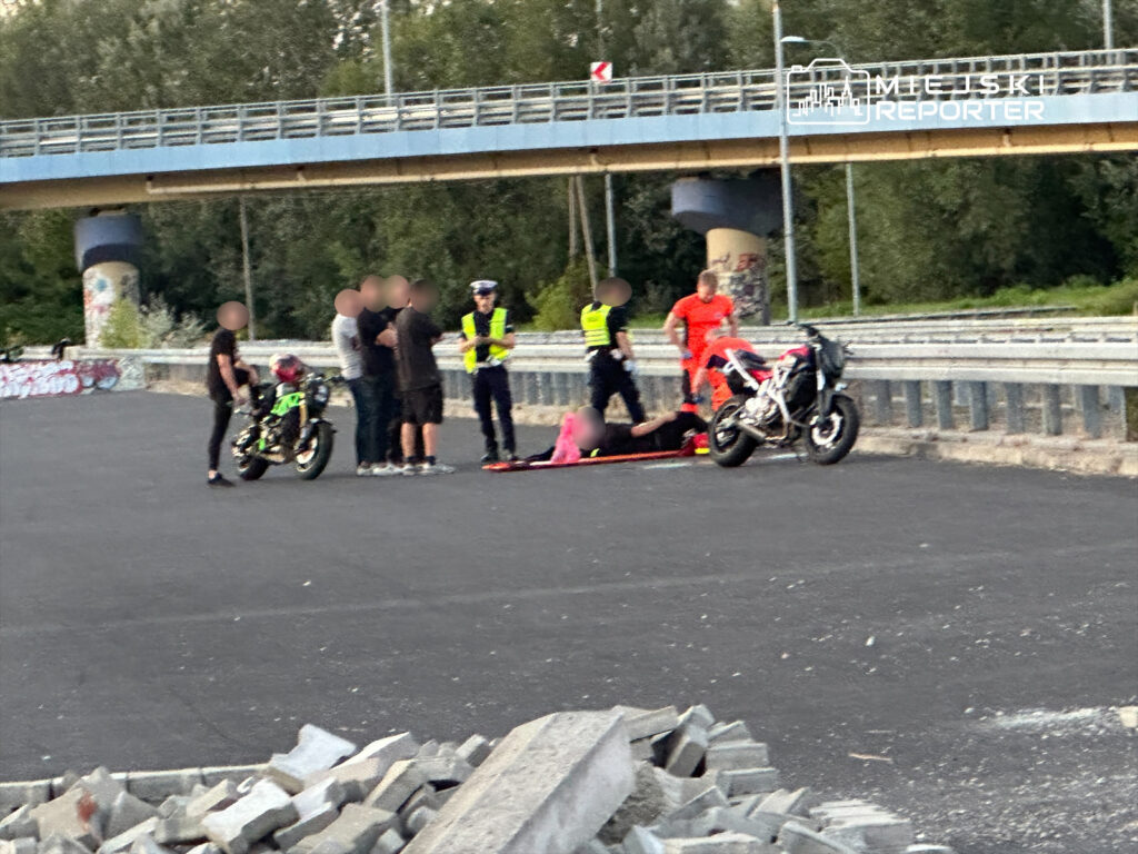 Grupa osób wokół leżącego mężczyzny na asfalcie, obok motocykli i funkcjonariuszy policji.