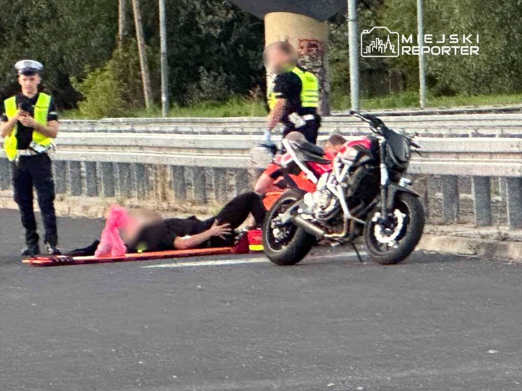 Mężczyzna leży na noszach, a obok niego stoi policjant i ratownik medyczny, obok zaparkowany motocykl.