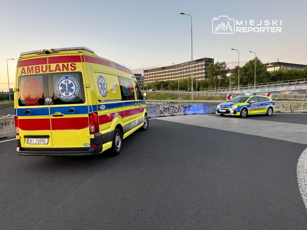 Ambulans i radiowóz policyjny stoją na zakręcie drogi, w tle nowoczesne budynki oraz graffiti na murze.