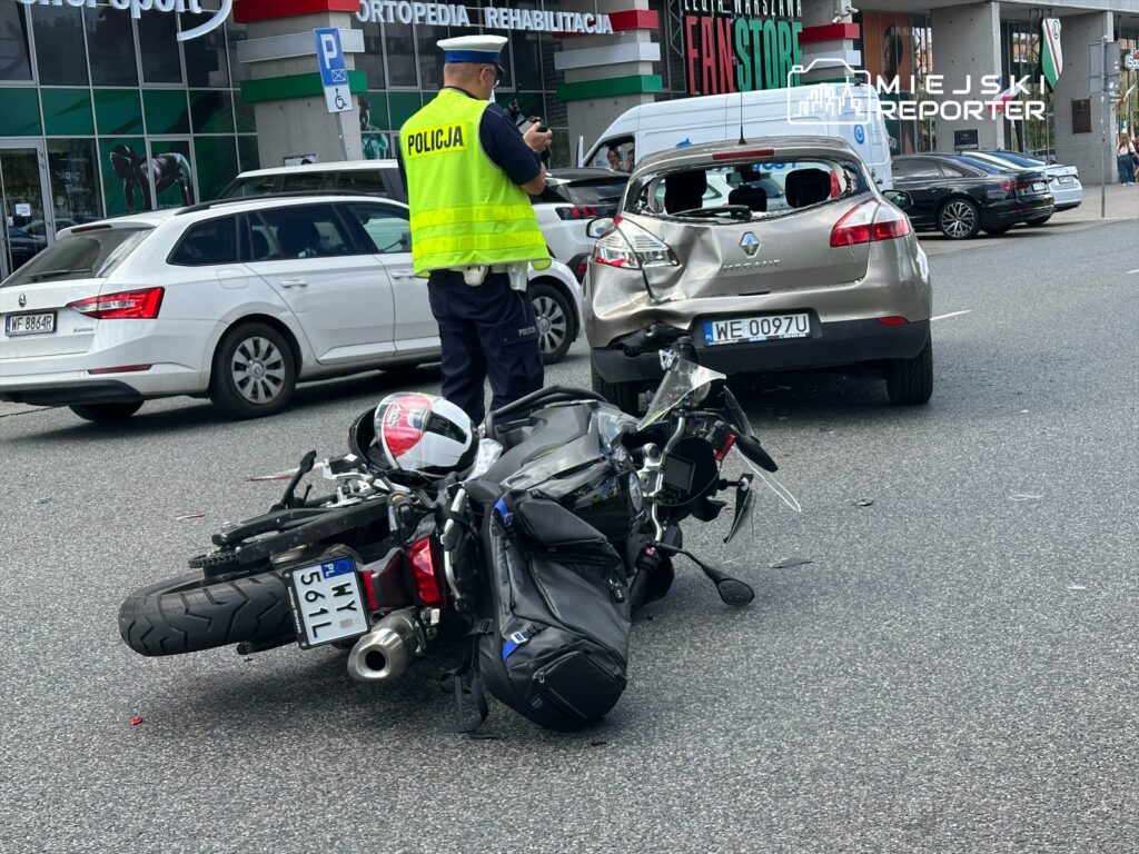 Policjant w kamizelce odblaskowej dokumentuje wypadek drogowy, obok leży przewrócony motocykl, a uszkodzony samochód stoi na jezdni.