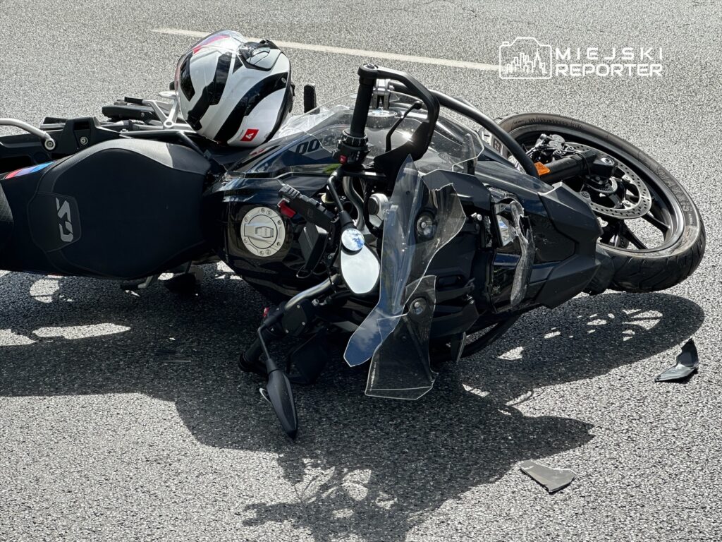 Motocykl leży na drodze, obok niego znajduje się kask. Na nawierzchni leżą fragmenty plastiku.