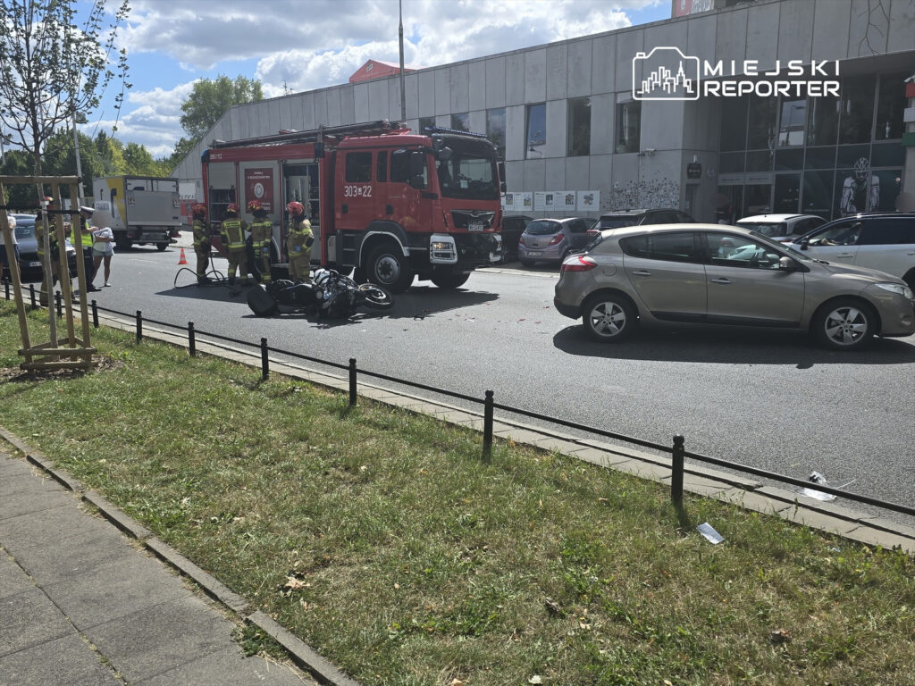 Grupa strażaków pracuje przy wypadku drogowym, obok leżącego motocykla, na ruchliwej ulicy.