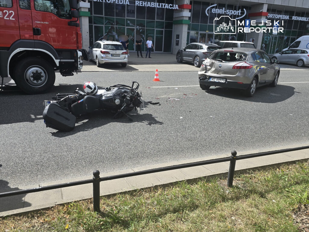Motocykl leży na jezdni obok uszkodzonego samochodu, a w tle widać budynek z reklamą oraz pojazdy zaparkowane na ulicy.