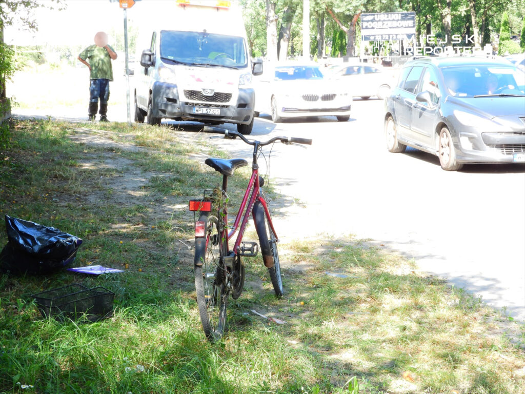 Rower w kolorze czerwonym zaparkowany na poboczu, obok czarnego worka na śmieci i złożonych papierów. W tle przejeżdżające samochody.