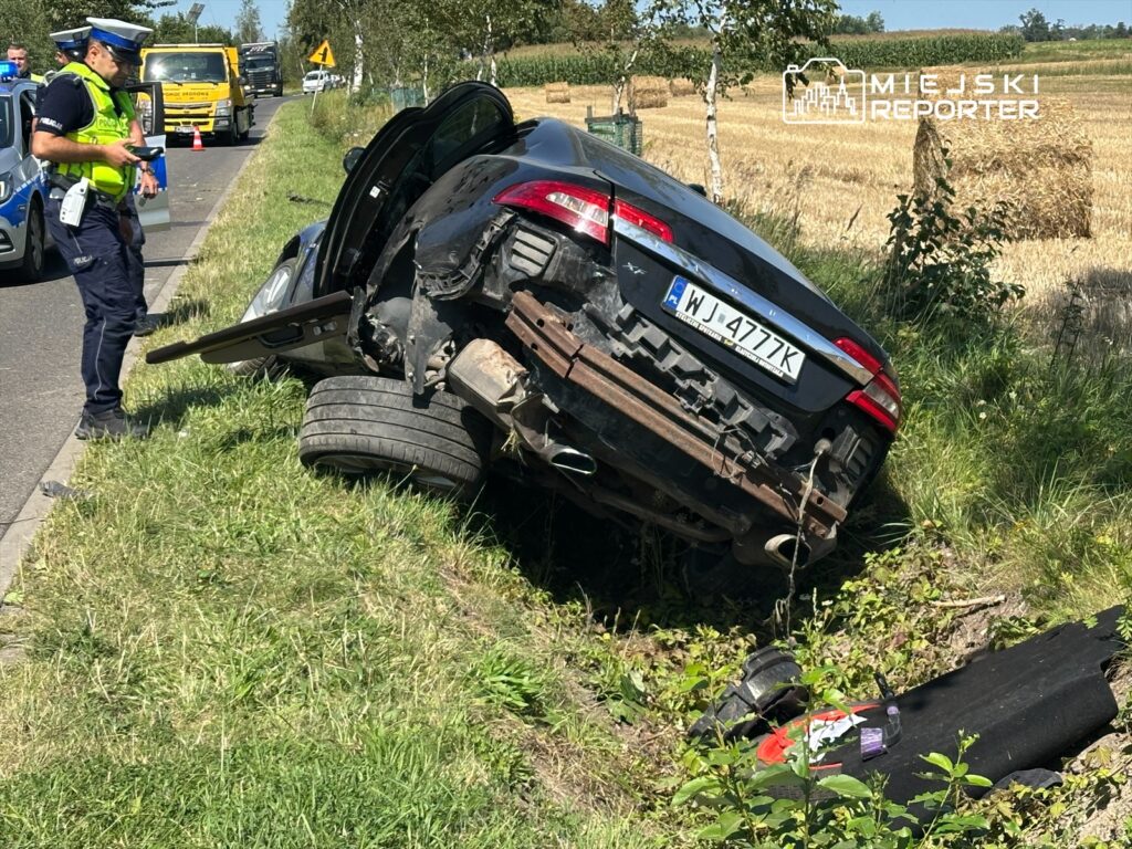 Radiowóz policyjny zatrzymany obok samochodu osobowego leżącego na boku w rowie, w otoczeniu pól.
