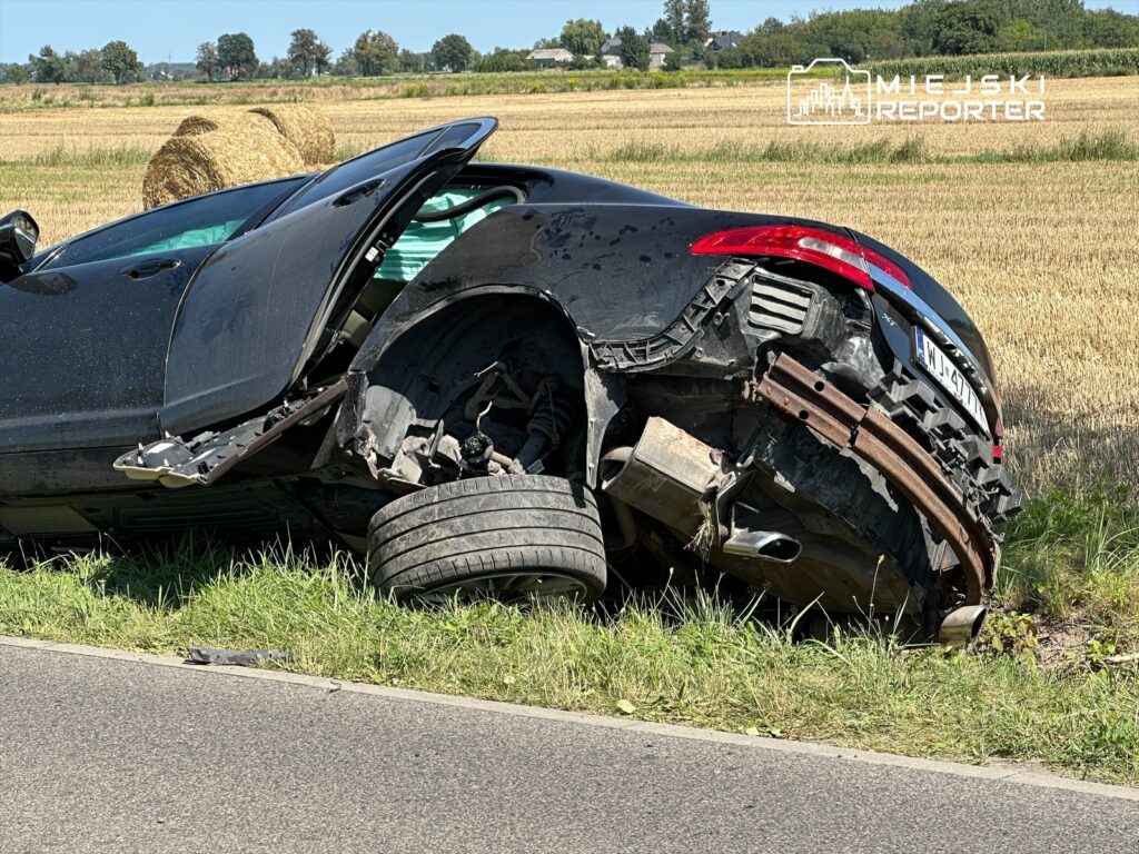 Czarny samochód sportowy leży na boku w zbożu, z uszkodzoną tylną osią i otwartymi drzwiami.
