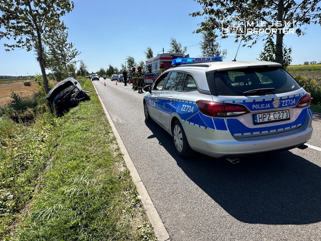 Na drodze stoi radiowóz policji z włączonymi sygnałami świetlnymi obok wraku samochodu w rowie.