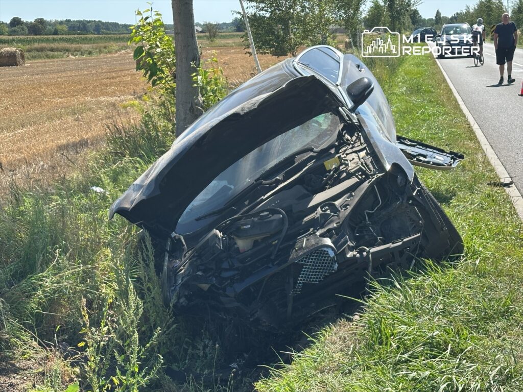 Czarny samochód osobowy leży na boku w zaroślach obok drogi, a w tle widać pola i przejeżdżających rowerzystów.
