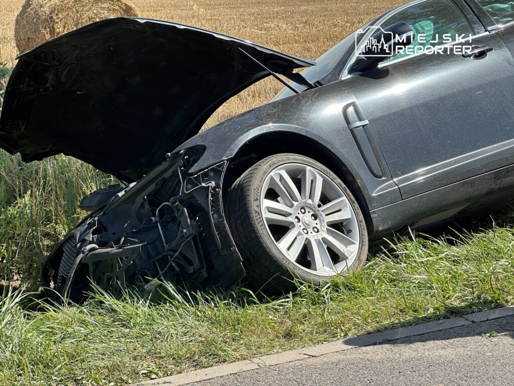 Uszkodzony samochód z otwartą maską stoi na poboczu drogi, obok pola z zebranym sianem.