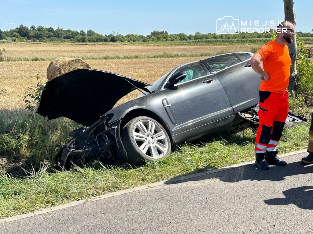 Samochód osobowy leży na poboczu drogi, a obok niego stoją mężczyzna w pomarańczowej koszulce i ratownik w odzieży ochronnej.