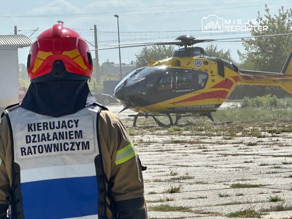 Kierujący działaniami ratowniczymi mężczyzna w hełmie obserwuje lądowanie helikoptera ratunkowego na placu.